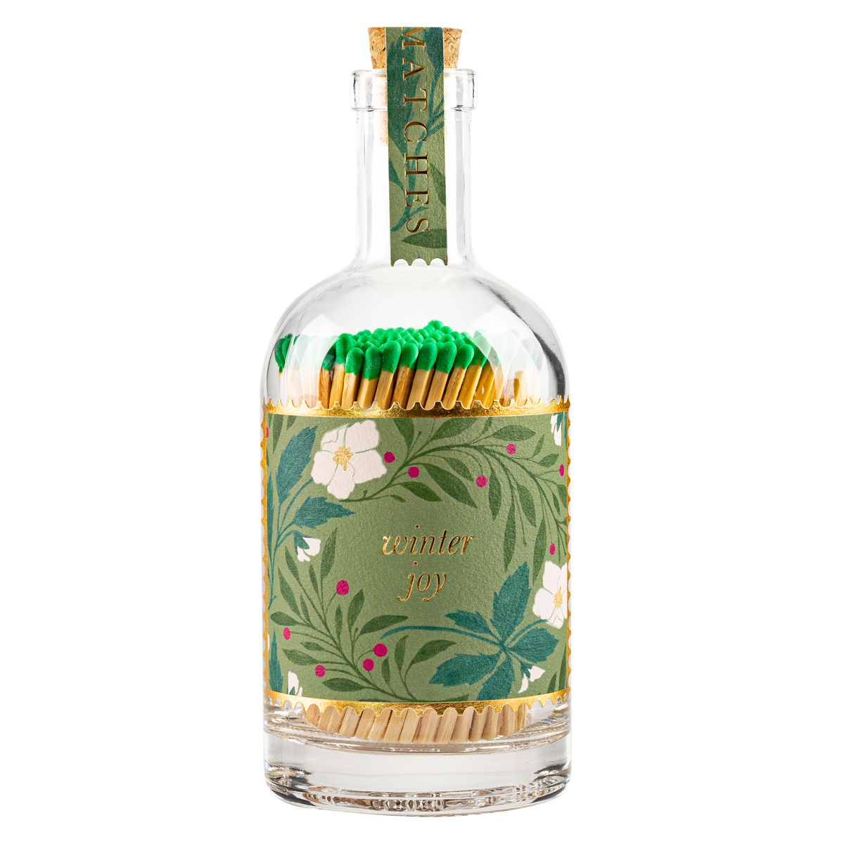A clear glass bottle filled with wooden matchsticks, wrapped in a green botanical paper sleeve with white flowers and gold lettering that reads 'winter joy.' The bottle is topped with a matching green paper neck wrap.