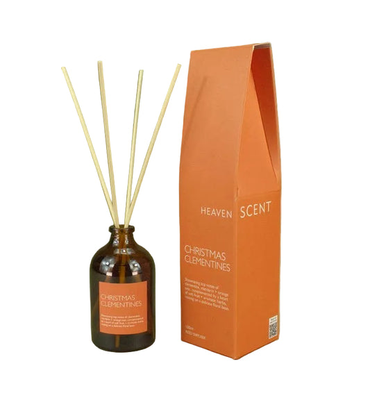 An amber glass reed diffuser bottle with four natural reeds and an orange label, standing next to its tall orange packaging box on a muted olive green background. The box is labeled 'HEAVEN SCENT' and 'CHRISTMAS ELEMPINES'.