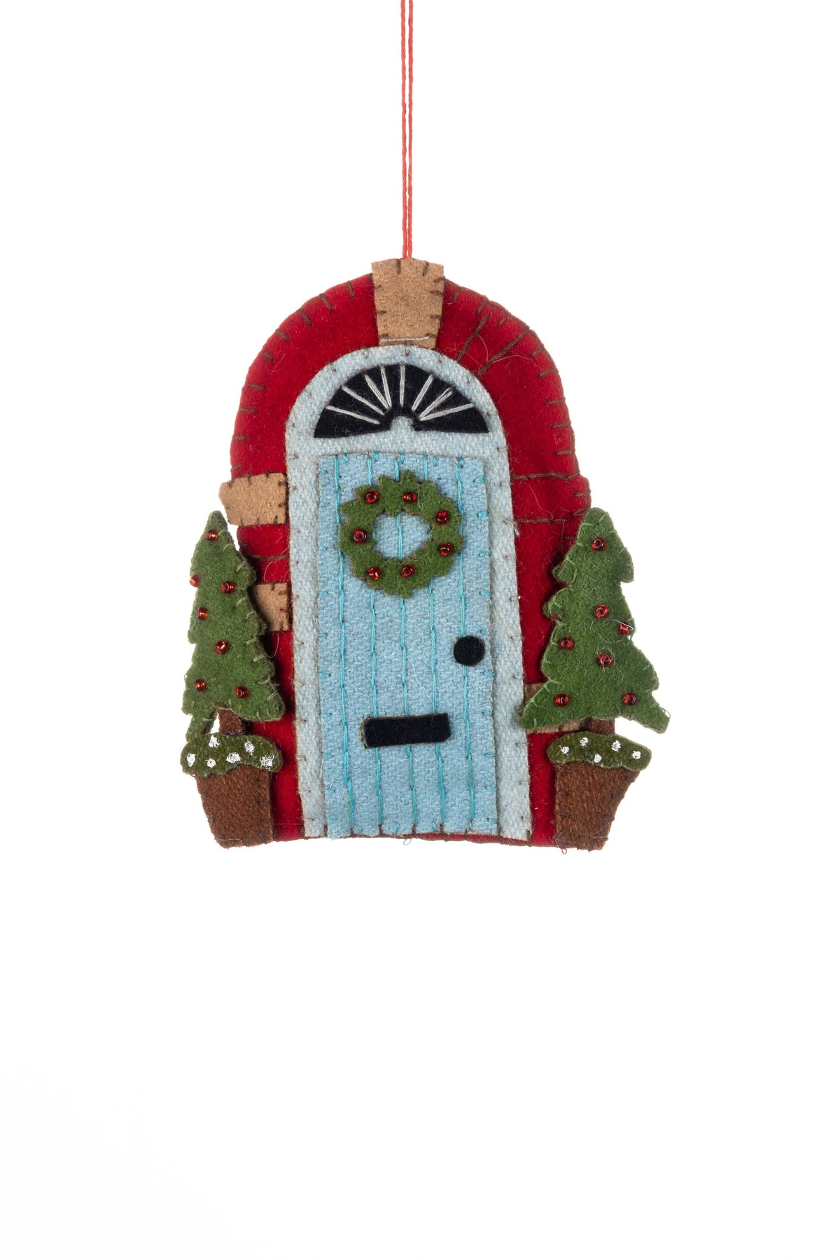 A felt Christmas ornament shaped like a small house with a red roof, light blue door, green wreath, and two green Christmas trees on either side, hanging by a thin red string against a white background.