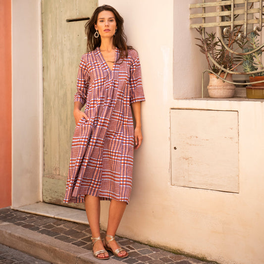 Woman in a plaid dress standing against a wall with a plant in the background