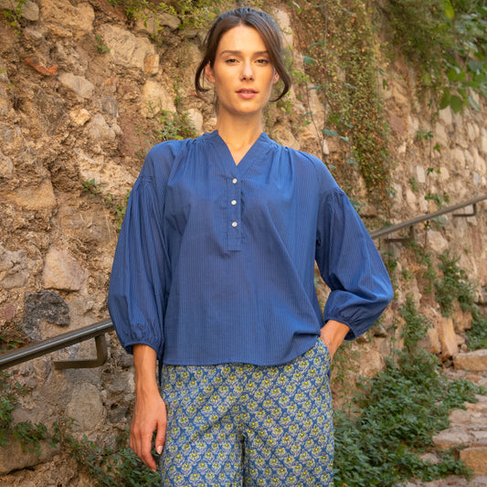 Woman wearing a blue blouse and patterned pants standing against a stone wall.