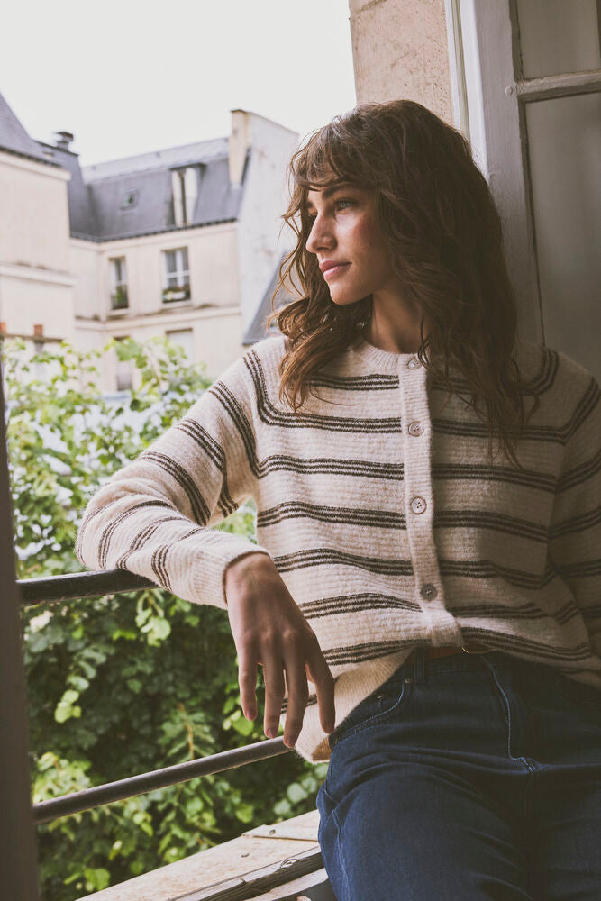 Fransa Kendra Cardigan in Oatmeal Stripe - A woman with wavy brown hair sits on a balcony wearing a cream-colored knit cardigan with thin dark brown horizontal stripes and dark blue jeans.