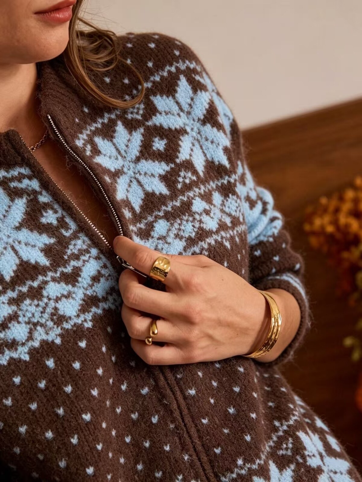 A close-up of a person wearing a brown and light blue patterned knit zip-up sweater, with gold rings and bracelets on their hand.