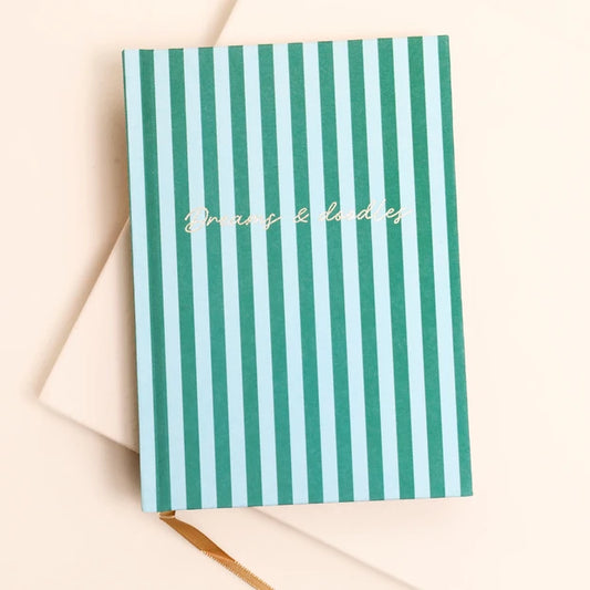 A hardcover notebook with vertical mint green and white stripes, featuring the phrase 'In any & ladies' in gold cursive script on the cover, styled on a cream envelope against a light beige background.