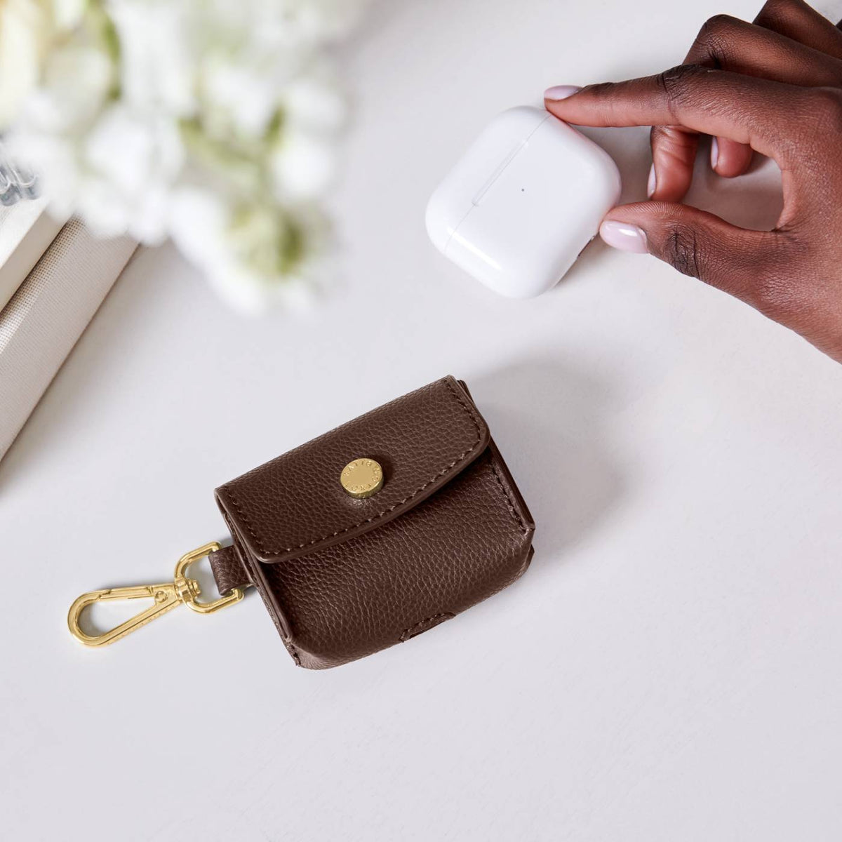 Katie Loxton Evie Clip On Airpod Case in Chocolate  - brown leather case with gold clasp on a white surface, next to a hand holding a white airpods case.