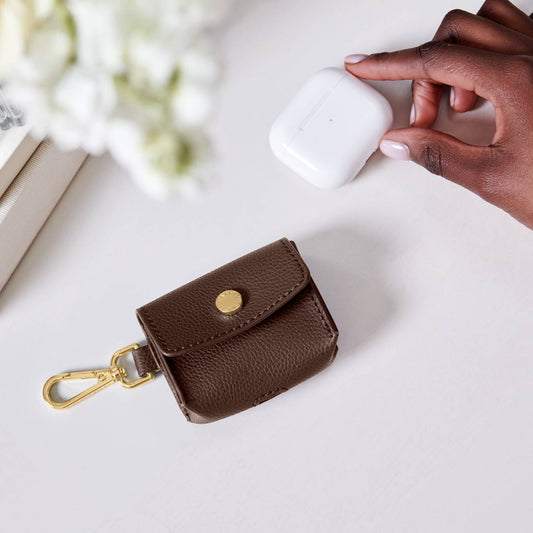 Katie Loxton Evie Clip On Airpod Case in Chocolate  - brown leather case with gold clasp on a white surface, next to a hand holding a white airpods case.