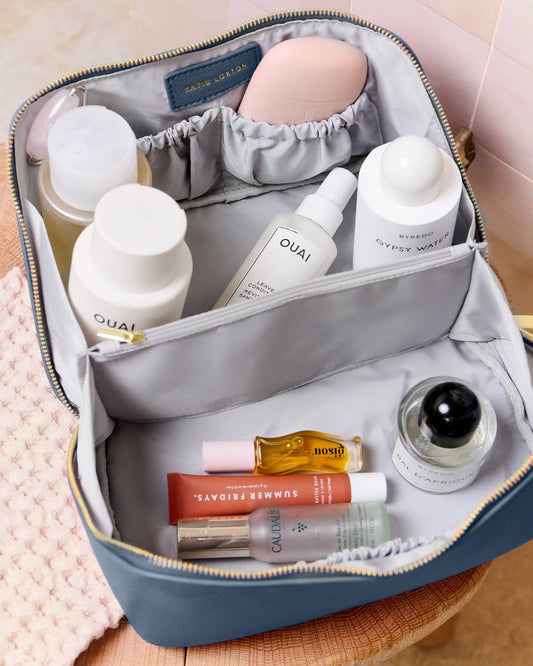 An open blue-gray toiletry bag with gold zipper trim, filled with skincare and beauty products. The interior organizer has elastic loops, side pockets, and a lower compartment holding bottles, tubes, and a small jar.