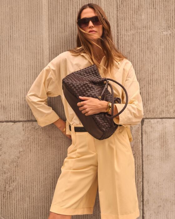 Woman holding a brown woven handbag against a textured wall.