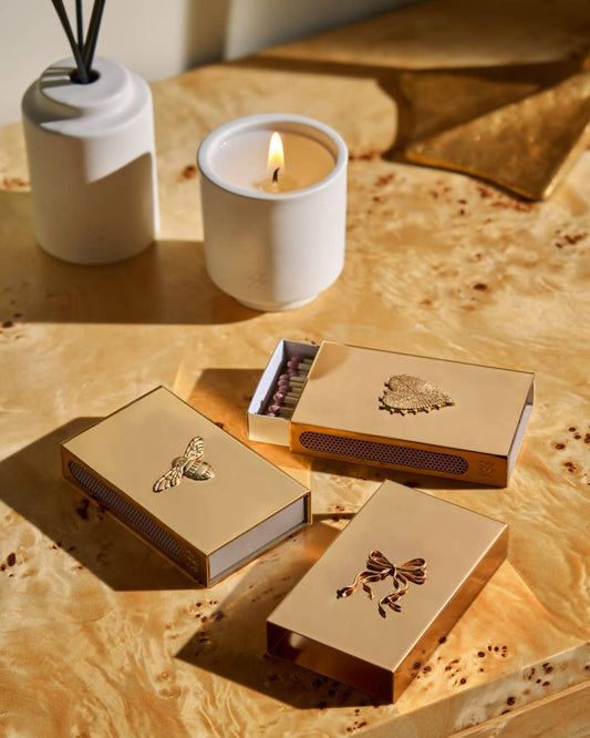 Three metallic gold matchboxes with decorative cutout designs are arranged on a warm marbled surface, alongside a lit white candle and a white diffuser in a styled indoor setting.