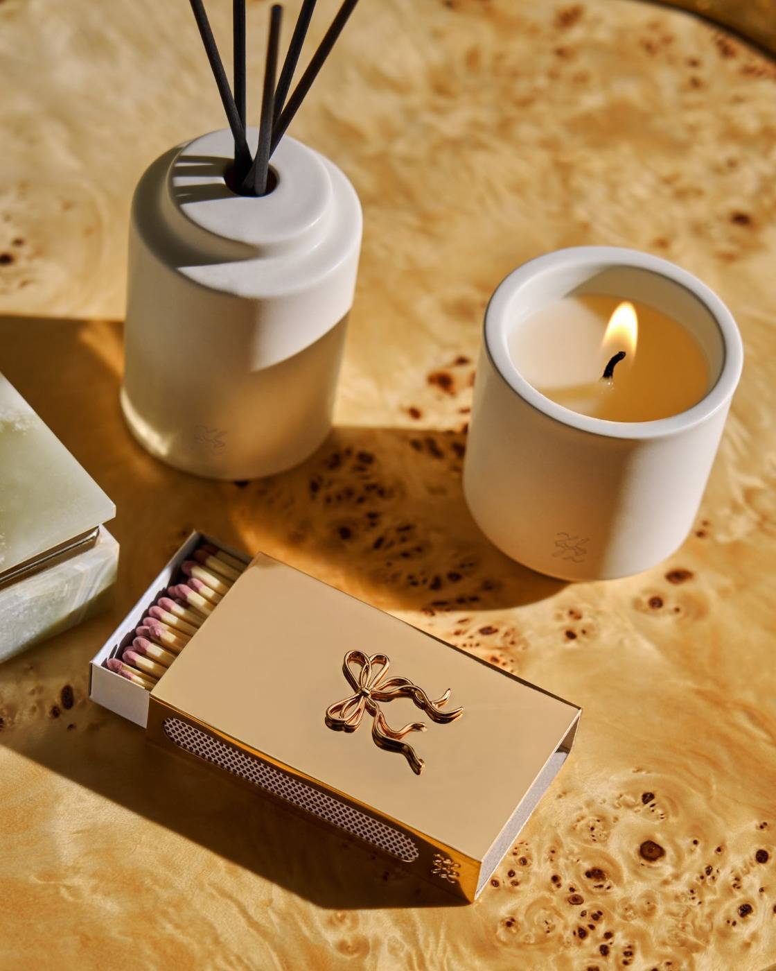 A styled product arrangement on a warm wood surface featuring a white ceramic candle with a lit flame, a white reed diffuser with black reeds, a gold-toned decorative matchbox, and part of a green soap bar.