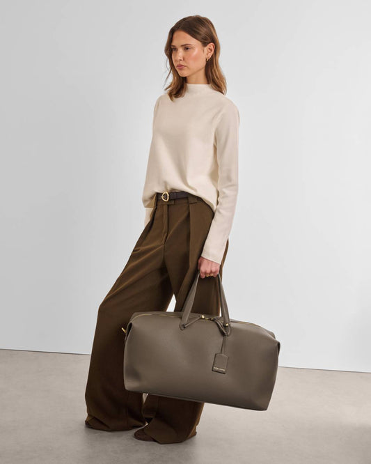 A woman stands in a studio wearing a cream mock-neck sweater, brown wide-leg trousers, and holding a large taupe leather duffel bag.
