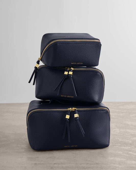Three stacked navy blue pebbled leather cosmetic pouches by Kate Leeton with gold zippers and tassel pulls, set against a neutral gray background.