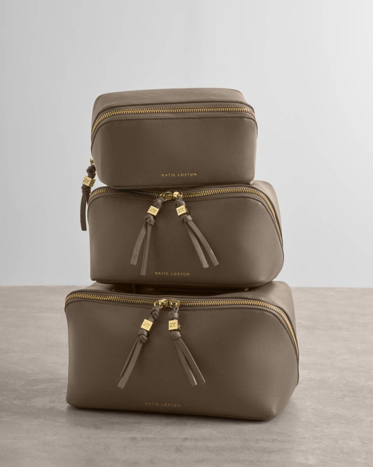 Three stacked rectangular makeup bags in a taupe-brown leather finish with gold-tone zippers and tassel pulls, photographed on a light gray studio background.