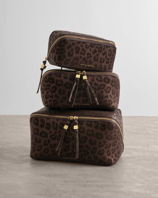 Three stacked Kate Spade cosmetic pouches in a dark brown leopard print fabric with gold-tone zippers and tassels, set against a neutral gray studio background.