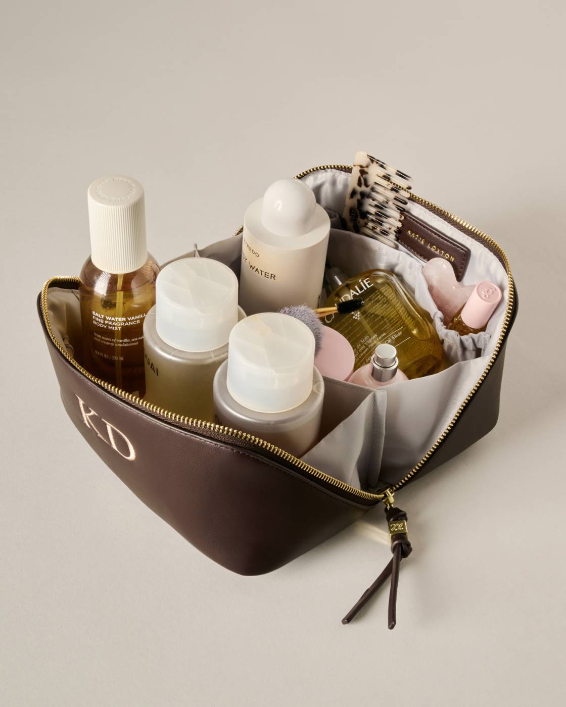 A brown zippered cosmetic bag with gold hardware and a monogrammed 'KD' on the front, shown open and filled with assorted skincare and perfume bottles on a light beige background.