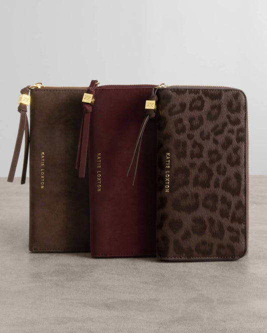 Three Kate Cotton zip-around wallets standing upright on a gray surface against a light gray background. The wallets are in three colors: dark brown, deep burgundy, and brown leopard print, each with a gold-tone zipper pull and gold brand lettering.
