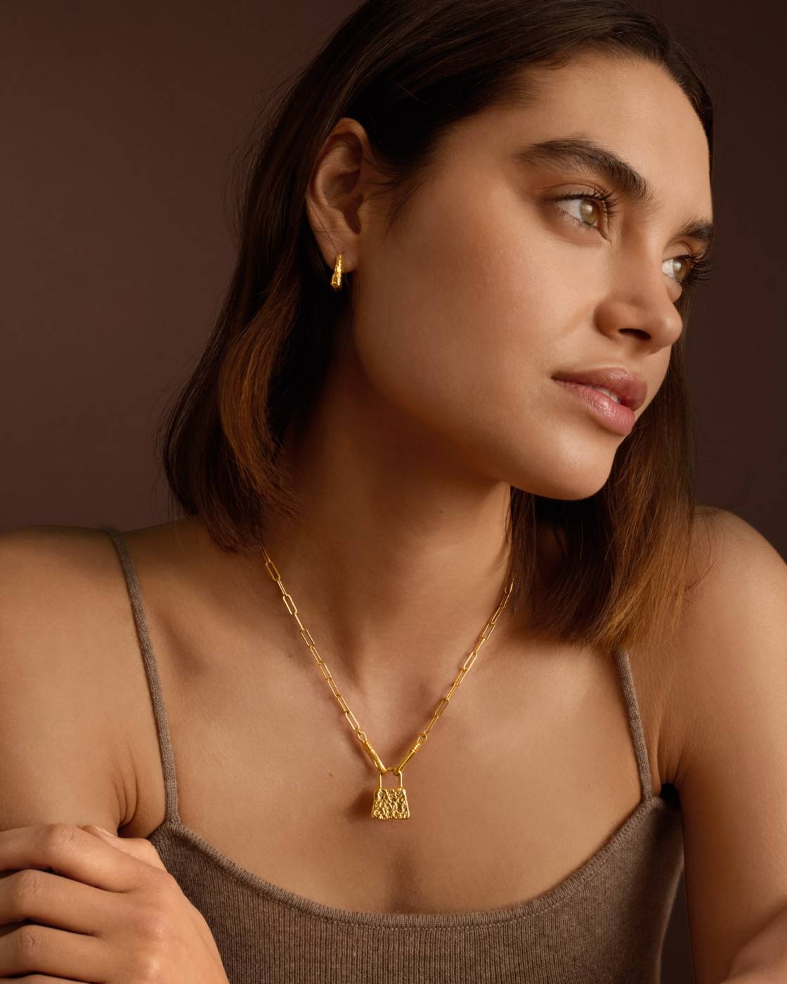 A woman wearing a delicate gold chain necklace with a small geometric pendant, styled with a matching small gold hoop earring, set against a warm brown background.