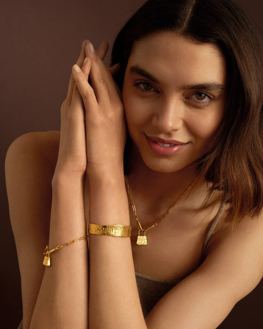 A close-up lifestyle shot of a woman wearing gold jewelry, featuring a textured gold bangle on her wrist and a matching gold pendant necklace, set against a dark brown background.