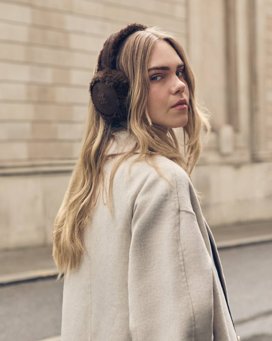 A woman with long blonde hair wearing dark brown fuzzy earmuffs and a light beige coat, photographed outdoors against a blurred stone building background.