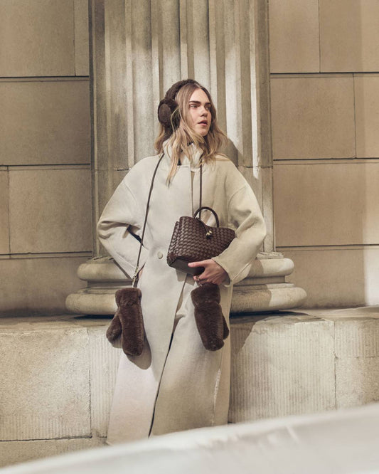 A woman stands on stone steps in front of a classical column, wearing a long cream coat, brown faux-fur earmuffs, matching brown mittens, and a brown woven handbag.