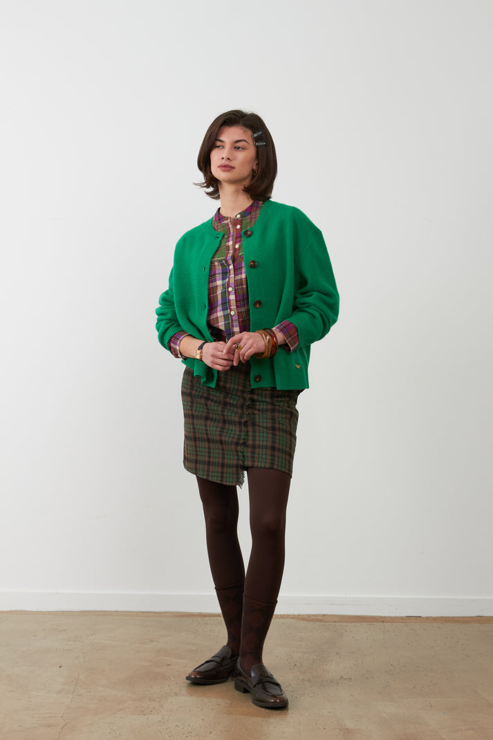 Orfeo Wayra Wool Cardigan in Green - a woman stands against a plain white wall wearing a bright green cardigan over a red and blue plaid shirt, paired with a green and red plaid mini skirt, dark brown tights, and black loafers.