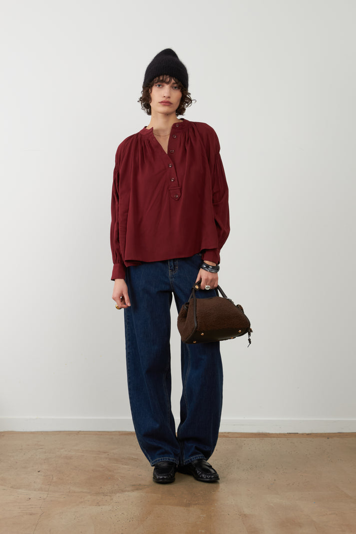 Orfeo Abby Shirt in Bordeaux - a model stands in a minimalist studio setting wearing a dark red long-sleeve blouse with a gathered neckline, dark blue wide-leg jeans, a black beanie, and holding a brown leather handbag.
