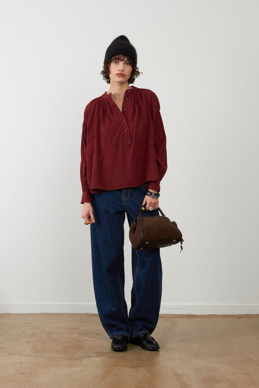 Orfeo Abby Shirt in Bordeaux - a model stands in a minimalist studio setting wearing a dark red long-sleeve blouse with a gathered neckline, dark blue wide-leg jeans, a black beanie, and holding a brown leather handbag.