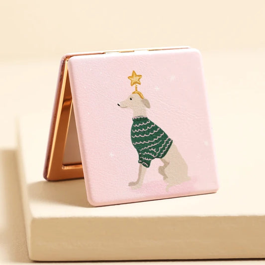 A square compact mirror with a light pink cover featuring an illustration of a dog wearing a green sweater and a gold star, shown standing open on a beige surface.