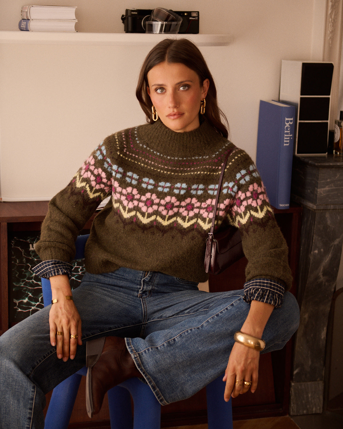 A woman sits on a blue stool wearing a dark olive green knit sweater with a colorful Fair Isle pattern across the chest and cuffs, paired with blue jeans and brown boots in a cozy indoor setting.