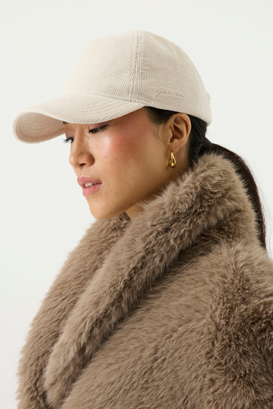 A woman in profile wearing a light beige corduroy baseball cap by Garcia and a large brown fur collar jacket against a plain light background.