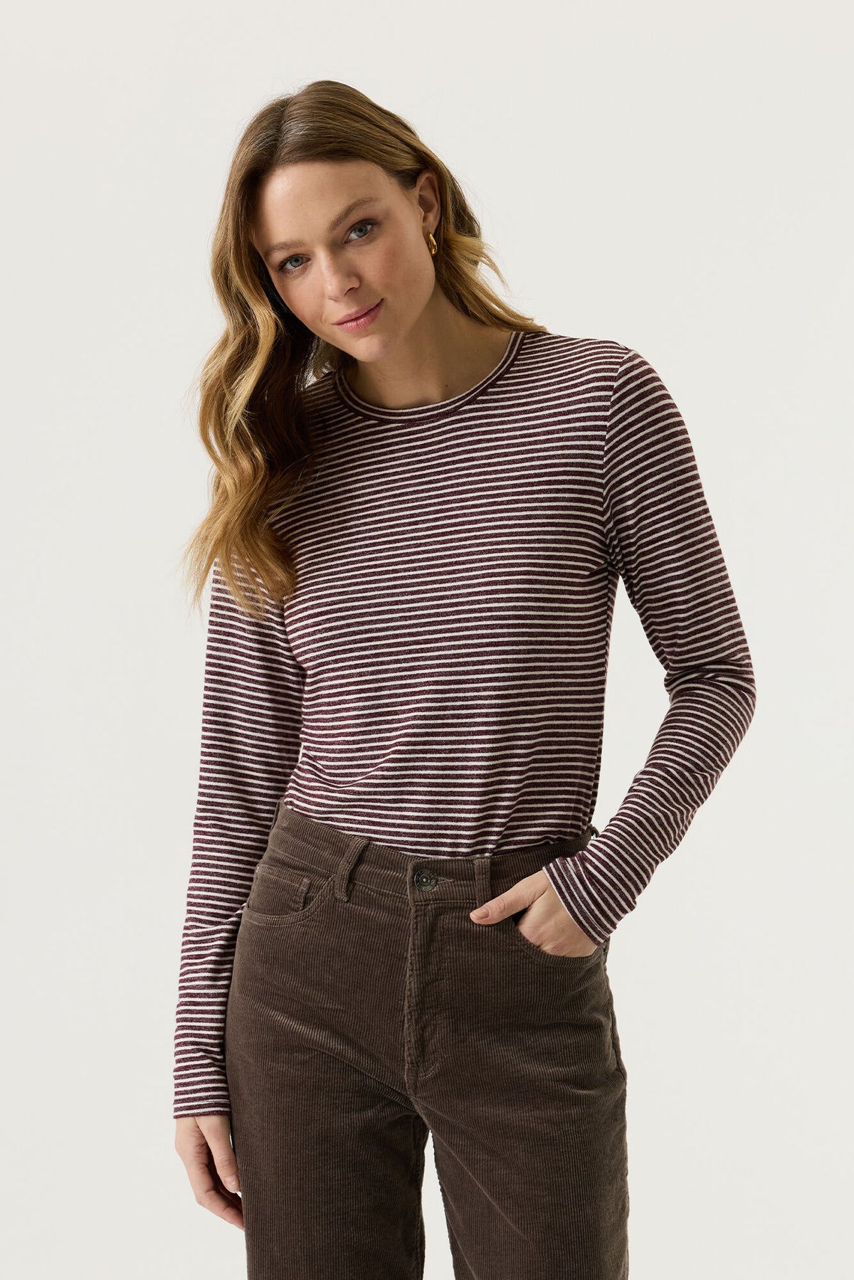 Garcia Long Sleeved Striped Tee in Rosewood - a woman modelling a long-sleeve crewneck top with thin dark brown and black horizontal stripes, paired with dark brown corduroy pants, against a light grey studio background.