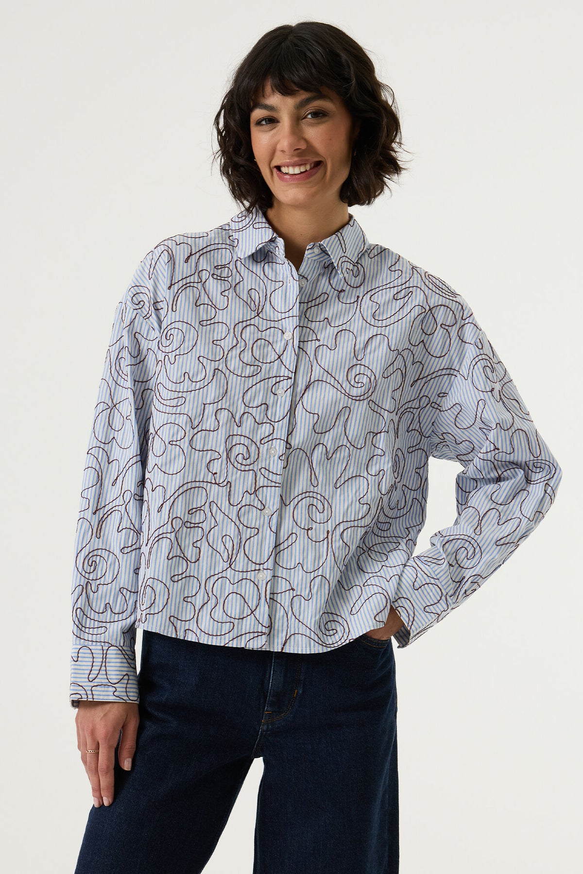 Garcia LS Striped Blouse with Embroidery in Breeze Blue - a woman wearing a light blue button-up shirt with a brown abstract line pattern, paired with dark blue jeans, against a plain light grey background.