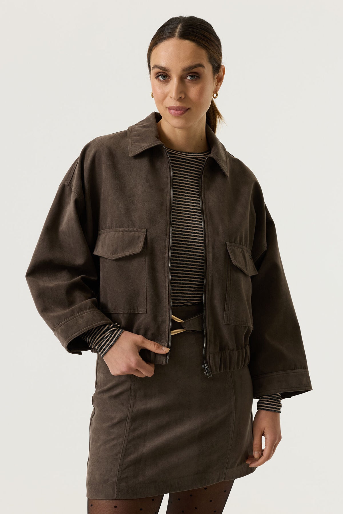 Garcia Graphite Grey Suede Jacket - A woman modelling a dark brown corduroy utility jacket with front flap pockets, worn over a black and white striped top and a matching brown skirt, against a light grey studio background.