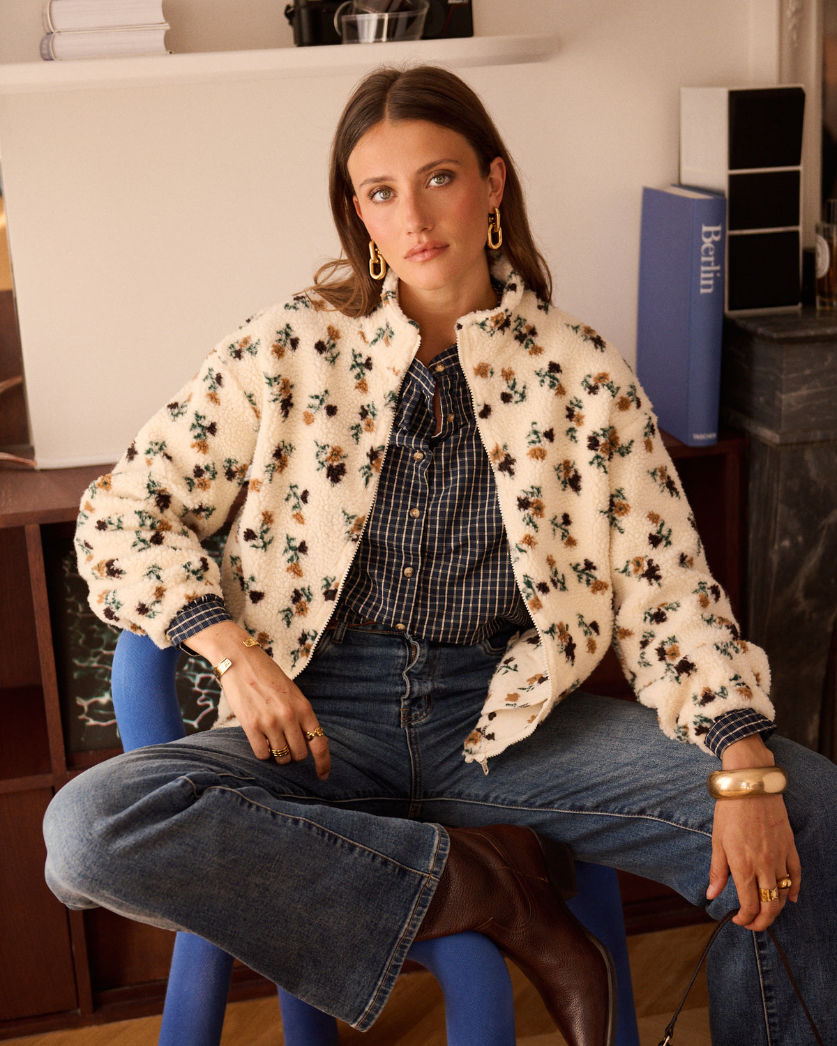A woman sits on a blue stool wearing a cream sherpa jacket with a brown and black floral pattern, a navy and white gingham shirt, blue jeans, and brown boots in an indoor room setting.