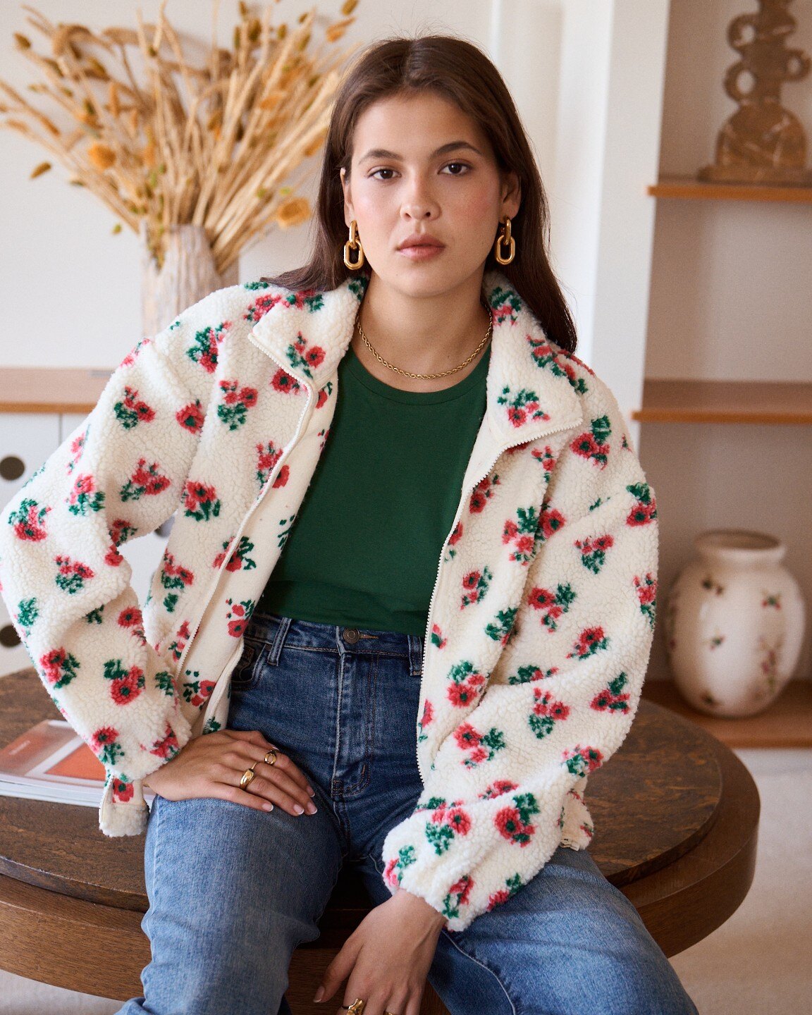 A woman wearing a cream-colored sherpa jacket with a red and green floral pattern, layered over a dark green t-shirt and blue jeans, sitting on a wooden table in a warm, styled room.